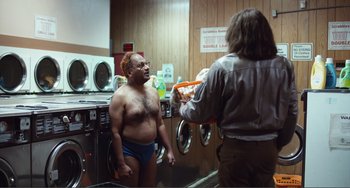 Movie still from “An Evening with Beverly Luff Linn” (2018), directed by Jim Hosking – A man in a blue swim trunks is talking to a woman in front of a washing machine; Wide shot, Low angle