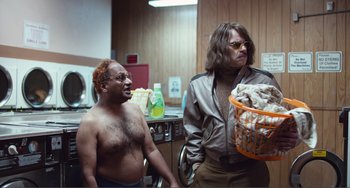 Movie still from “An Evening with Beverly Luff Linn” (2018), directed by Jim Hosking – Two men are standing in a kitchen and one is holding a basket of laundry; Medium shot, Over the shoulder angle