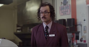 Movie still from “An Evening with Beverly Luff Linn” (2018), directed by Jim Hosking – A man in a suit and tie standing in front of a counter; Medium shot, Low angle
