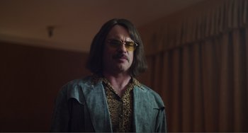 Movie still from “An Evening with Beverly Luff Linn” (2018), directed by Jim Hosking – A man with long brown hair and a leopard print shirt; Medium shot, Over the shoulder angle