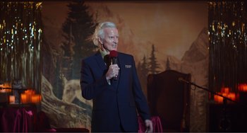 Movie still from “An Evening with Beverly Luff Linn” (2018), directed by Jim Hosking – A man in a blue suit holding a microphone; Medium shot, Over the shoulder angle