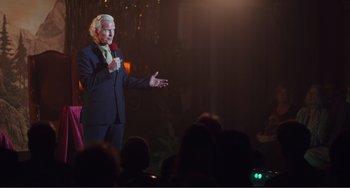 Movie still from “An Evening with Beverly Luff Linn” (2018), directed by Jim Hosking – An older man in a suit is speaking to a crowd; Medium shot, Over the shoulder angle
