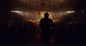 Movie still from “An Evening with Beverly Luff Linn” (2018), directed by Jim Hosking – A man standing in front of an audience in a dark room; Wide shot, Over the shoulder angle