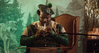 Movie still from “An Evening with Beverly Luff Linn” (2018), directed by Jim Hosking – A man in a green outfit is playing a flute; Medium shot, High angle