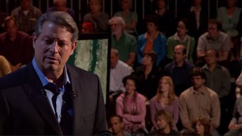Movie still from “An Inconvenient Truth” (2006), directed by Davis Guggenheim – A crowd of people sitting and standing in front of a man in a suit; Medium shot, Over the shoulder angle