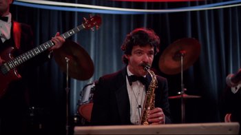 Movie still from “An Officer and a Gentleman” (1982), directed by Taylor Hackford – A man in a tuxedo playing a saxophone in front of other instruments; Medium shot, High angle