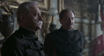 Movie still from “An Officer and a Spy” (2019), directed by Roman Polanski – Two older men in black jackets standing next to each other in a room; Medium shot, Low angle