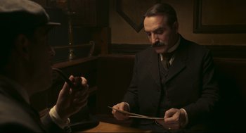 Movie still from “An Officer and a Spy” (2019), directed by Roman Polanski – An older man sitting at a table with a cell phone; Medium shot, Over the shoulder angle