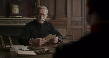 Movie still from “An Officer and a Spy” (2019), directed by Roman Polanski – An older man sitting at a table with a piece of paper in front of him; Medium shot, Over the shoulder angle