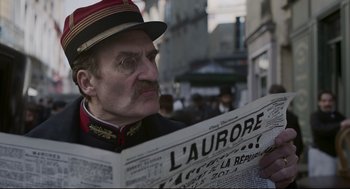 Movie still from “An Officer and a Spy” (2019), directed by Roman Polanski – A man wearing a hat and holding a newspaper in his hands; Close Up shot, Low angle