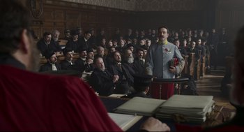 Movie still from “An Officer and a Spy” (2019), directed by Roman Polanski – A man in a crowd of people sitting in front of a microphone; Wide shot, Low angle