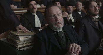 Movie still from “An Officer and a Spy” (2019), directed by Roman Polanski – An old man with a beard and glasses sitting in front of a group of men; Medium shot, Over the shoulder angle