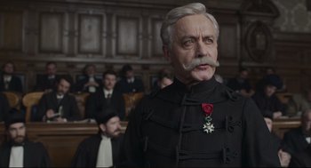 Movie still from “An Officer and a Spy” (2019), directed by Roman Polanski – An older man in a black suit with a medal on his chest; Close Up shot, Over the shoulder angle