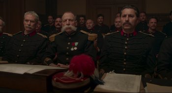 Movie still from “An Officer and a Spy” (2019), directed by Roman Polanski – A group of men in military uniforms sitting in front of a red hat; Medium shot, Low angle