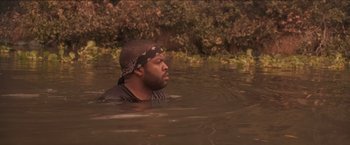 Movie still from “Anaconda” (1997), directed by Luis Llosa – A man in a black shirt and a black hat is in the water; Medium shot, High angle