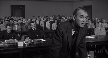 Movie still from “Anatomy of a Murder” (1959), directed by Otto Preminger – A man in a suit and tie in front of a group of people; Medium shot, Over the shoulder angle