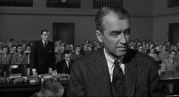 Movie still from “Anatomy of a Murder” (1959), directed by Otto Preminger – A man in a suit and tie in front of a group of people; Medium shot, Over the shoulder angle