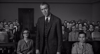 Movie still from “Anatomy of a Murder” (1959), directed by Otto Preminger – A man standing in front of a group of people in a courtroom; Medium shot, Over the shoulder angle