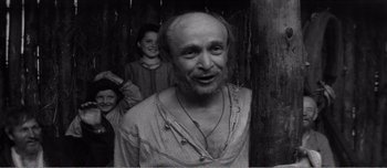 Movie still from “Andrei Rublev” (1966), directed by Andrei Tarkovsky – An old man with a beard and a long beard is smiling for the camera while two other people are standing behind him; Medium shot, Over the shoulder angle