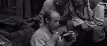 Movie still from “Andrei Rublev” (1966), directed by Andrei Tarkovsky – A black and white photo of a man eating something; Medium shot, High angle