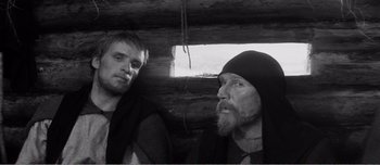 Movie still from “Andrei Rublev” (1966), directed by Andrei Tarkovsky – Two men are standing next to each other in front of a log cabin; Medium shot, Low angle