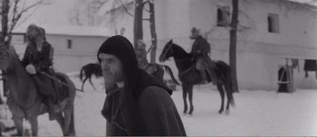 Movie still from “Andrei Rublev” (1966), directed by Andrei Tarkovsky – A man with a long beard and a hooded jacket is walking in the snow with two men on horses; Medium shot, High angle