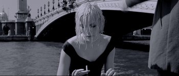 Movie still from “Angel-A” (2005), directed by Luc Besson – A black and white photo of a woman smoking a cigarette in front of a bridge; Medium shot, Low angle