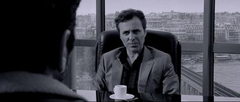 Movie still from “Angel-A” (2005), directed by Luc Besson – A man sitting at a table with a cup of coffee; Medium shot, Over the shoulder angle