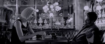 Movie still from “Angel-A” (2005), directed by Luc Besson – A woman sitting at a table in front of a cake on a plate; Medium shot, Over the shoulder angle