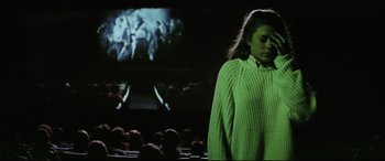 Movie still from “Anguish” (1987), directed by Bigas Luna – A woman standing in front of an audience wearing a green sweater; Medium shot, Low angle