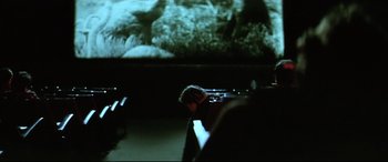 Movie still from “Anguish” (1987), directed by Bigas Luna – A person sitting in a theater with a projector screen in the background; Wide shot, Over the shoulder angle