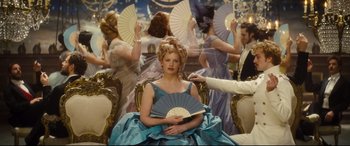 Movie still from “Anna Karenina” (2012), directed by Joe Wright – A woman sitting in a chair holding a fan in front of a group of people; Medium shot, Over the shoulder angle