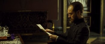 Movie still from “Anna Karenina” (2012), directed by Joe Wright – A man sitting at a table looking at a piece of paper; Medium shot, Over the shoulder angle