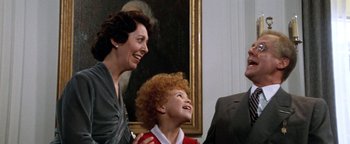 Movie still from “Annie” (1982), directed by John Huston – Two people and a child smiling at each other in front of a painting; Medium shot, Over the shoulder angle