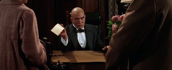 Movie still from “Annie” (1982), directed by John Huston – A man sitting at a table holding up a piece of paper; Close Up shot, Over the shoulder angle