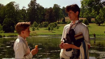 Movie still from “Another Country” (1984), directed by Marek Kanievska – A man standing next to a young boy near a body of water; Medium shot, Over the shoulder angle