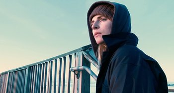Movie still from “Another Earth” (2011), directed by Mike Cahill – A woman wearing a hooded jacket standing in front of a fence; Close Up shot, Low angle