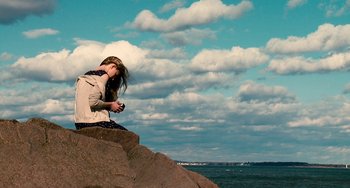 Movie still from “Another Earth” (2011), directed by Mike Cahill – A woman sitting on top of a rock near the ocean; Wide shot, High angle