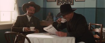 Movie still from “Appaloosa” (2008), directed by Ed Harris – Two men sitting at a table with a newspaper and two cups; Medium shot, Over the shoulder angle