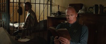 Movie still from “Appaloosa” (2008), directed by Ed Harris – An older man reading a book in front of an iron gate; Medium shot, Low angle