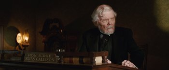 Movie still from “Appaloosa” (2008), directed by Ed Harris – An older man sitting at a desk in a suit and tie; Medium shot, Low angle