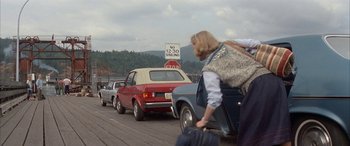 Movie still from “April Fool's Day” (1986), directed by Fred Walton – A woman pulling a suitcase behind a car on a road; Wide shot, Over the shoulder angle