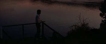 Movie still from “April Fool's Day” (1986), directed by Fred Walton – A woman standing on a pier near a body of water at night; Wide shot, Over the shoulder angle