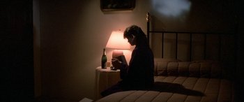 Movie still from “April Fool's Day” (1986), directed by Fred Walton – A person sitting on a bed reading a book next to a lamp; Medium shot, High angle