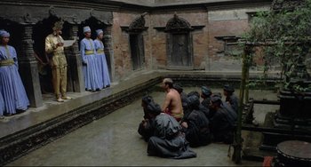Movie still from “Arabian Nights” (1974), directed by Pier Paolo Pasolini – A group of men sitting in a courtyard with a woman standing in front of them; Wide shot, High angle