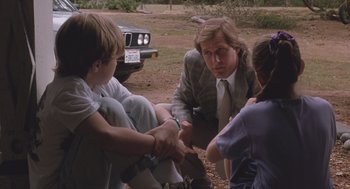 Movie still from “Arachnophobia” (1990), directed by Frank Marshall – A group of people sitting on the ground with a car in the background; Medium shot, Over the shoulder angle