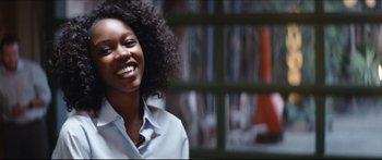 Movie still from “Archenemy” (2020), directed by Adam Egypt Mortimer – A woman smiles brightly while standing in front of a window; Close Up shot, Over the shoulder angle