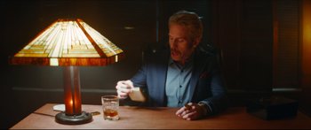 Movie still from “Archenemy” (2020), directed by Adam Egypt Mortimer – A man sitting at a table in front of a glass of whiskey; Medium shot, Low angle