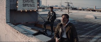 Movie still from “Archenemy” (2020), directed by Adam Egypt Mortimer – A man sitting on top of a bench next to another man holding a skateboard; Wide shot, Low angle