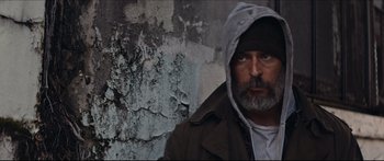 Movie still from “Archenemy” (2020), directed by Adam Egypt Mortimer – A man wearing a hooded jacket and glasses in front of a wall; Close Up shot, Low angle